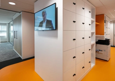 Moderne kantoorruimte aan de Karel Doormanweg met witte lockers, een tv-scherm en een printer op een feloranje vloer.